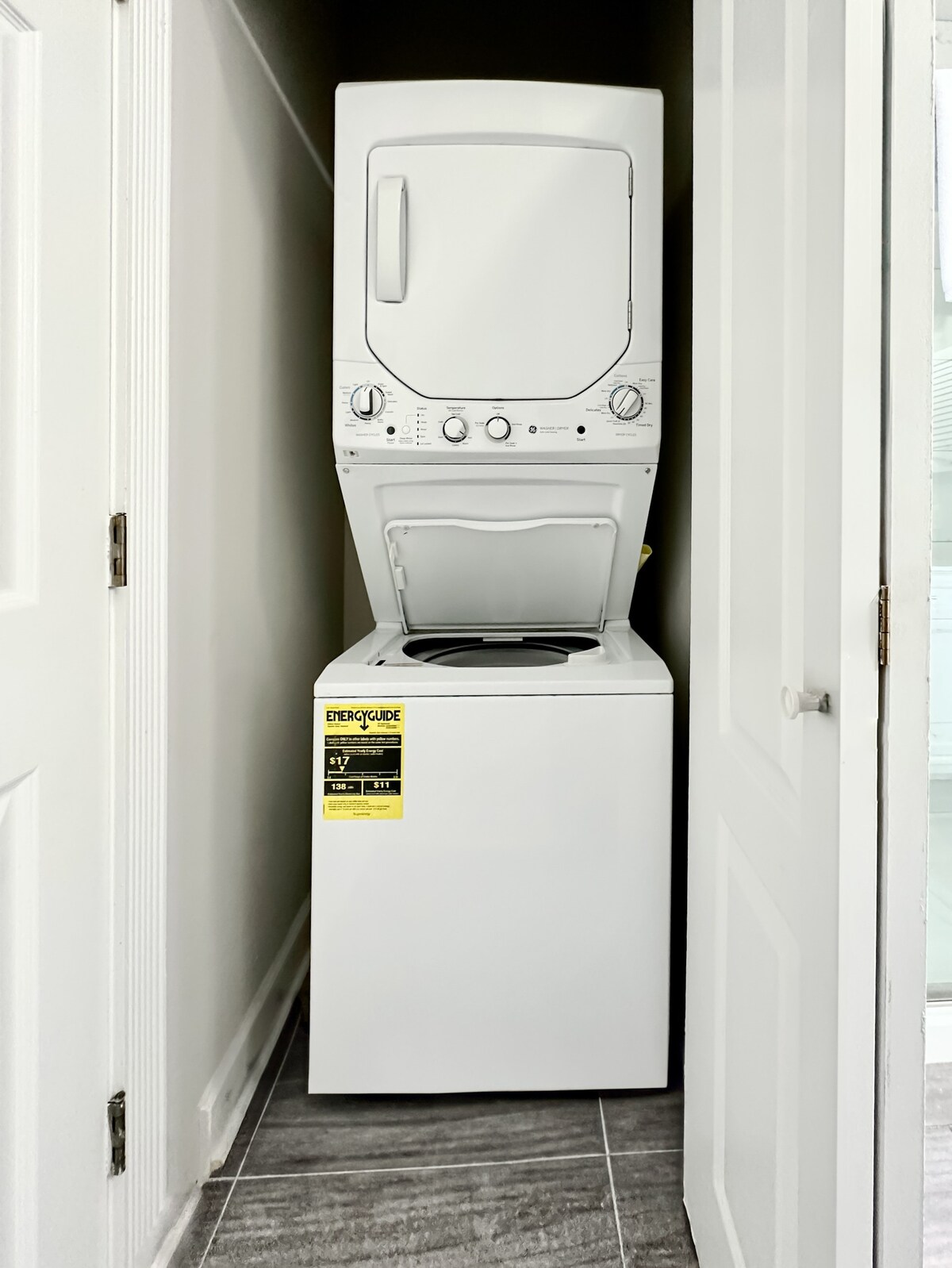 A stacked washer and dryer set is located in a designated laundry space, positioned between two white doors. The appliances are modern and energy-efficient, featuring visible dials and controls on the front. The floor is covered with gray tiles, contributing to the overall clean appearance.