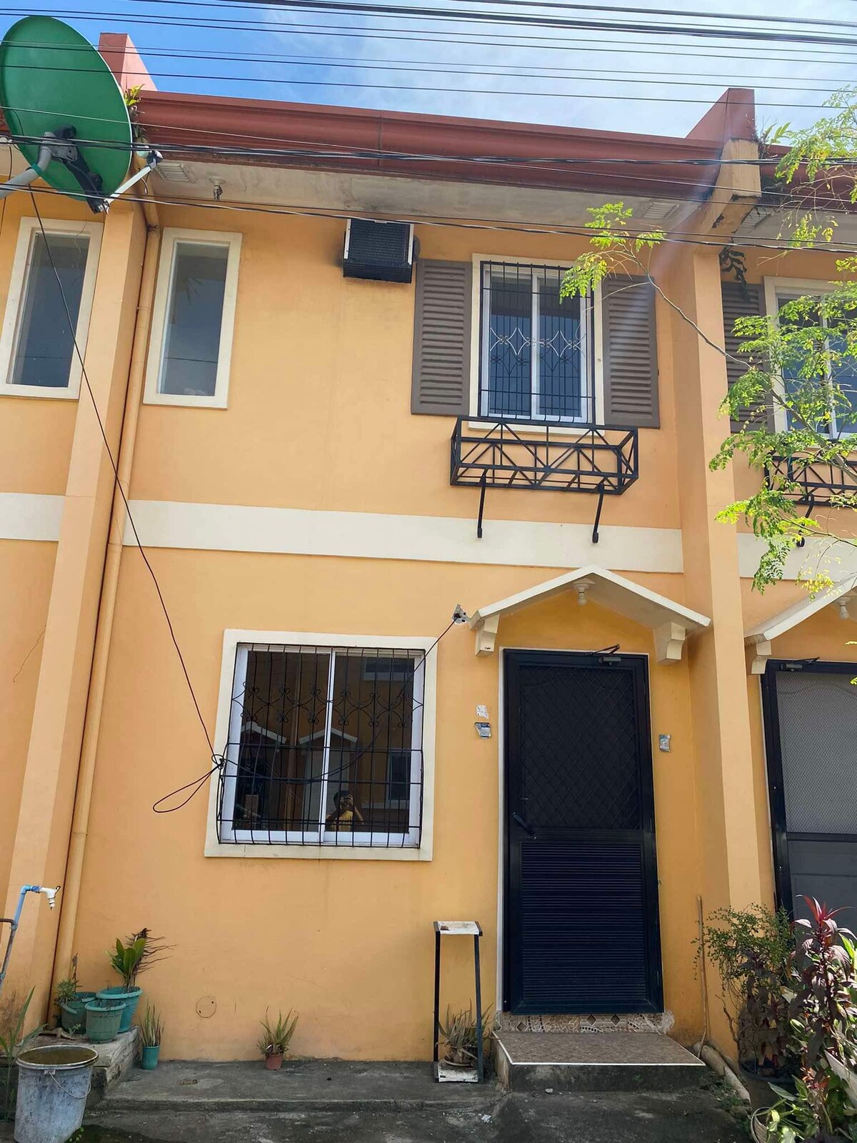 A charming two-story townhouse features a vibrant yellow exterior. Decorative shutters frame a window above the front door, while a small balcony adds character. A satellite dish is mounted on the roof, and plants are positioned along the ground, enhancing the welcoming appearance.