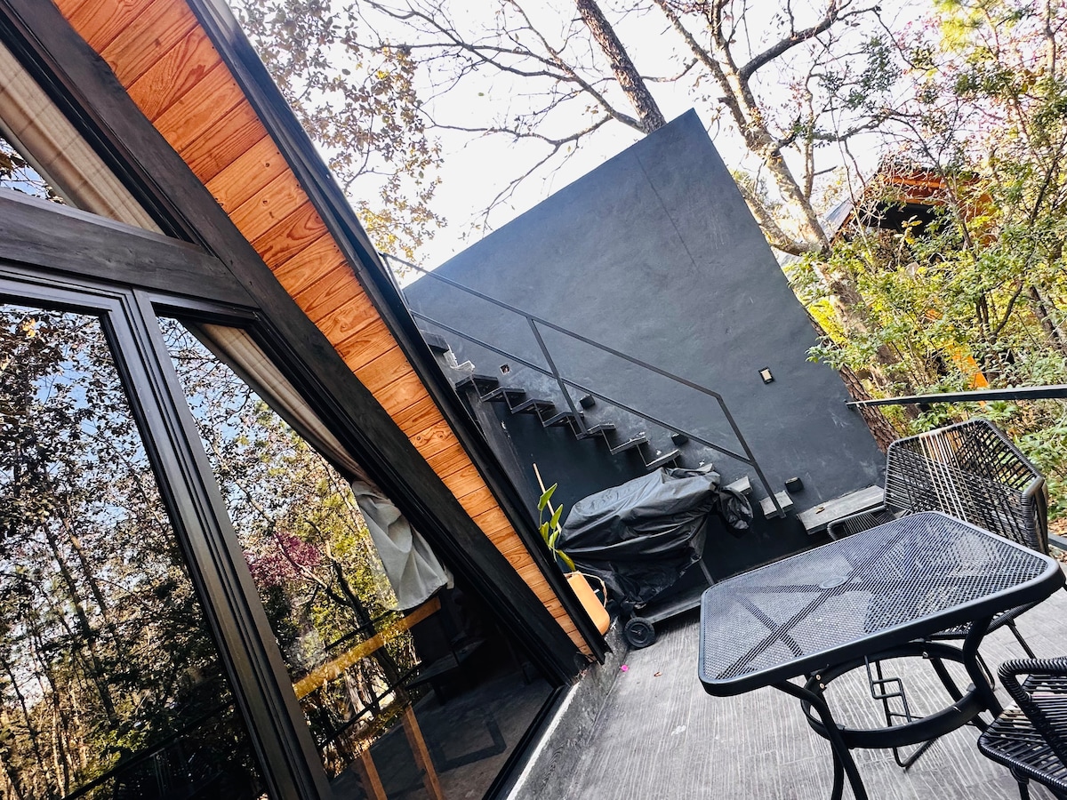 A contemporary terrace is highlighted, featuring a black metal staircase leading to the upper level. Two black chairs and a small table are positioned on the deck, with a grill situated nearby. Lush greenery surrounds the outdoor space, enhancing the natural setting.