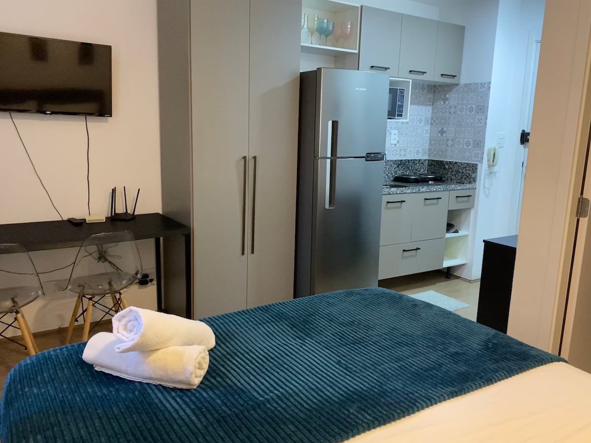 A studio living area is shown, featuring a spacious queen bed with folded towels on top. A modern kitchen is visible in the background, complete with a large refrigerator, sleek cabinets, and decorative wall tiles. A flat-screen TV is mounted on the wall.