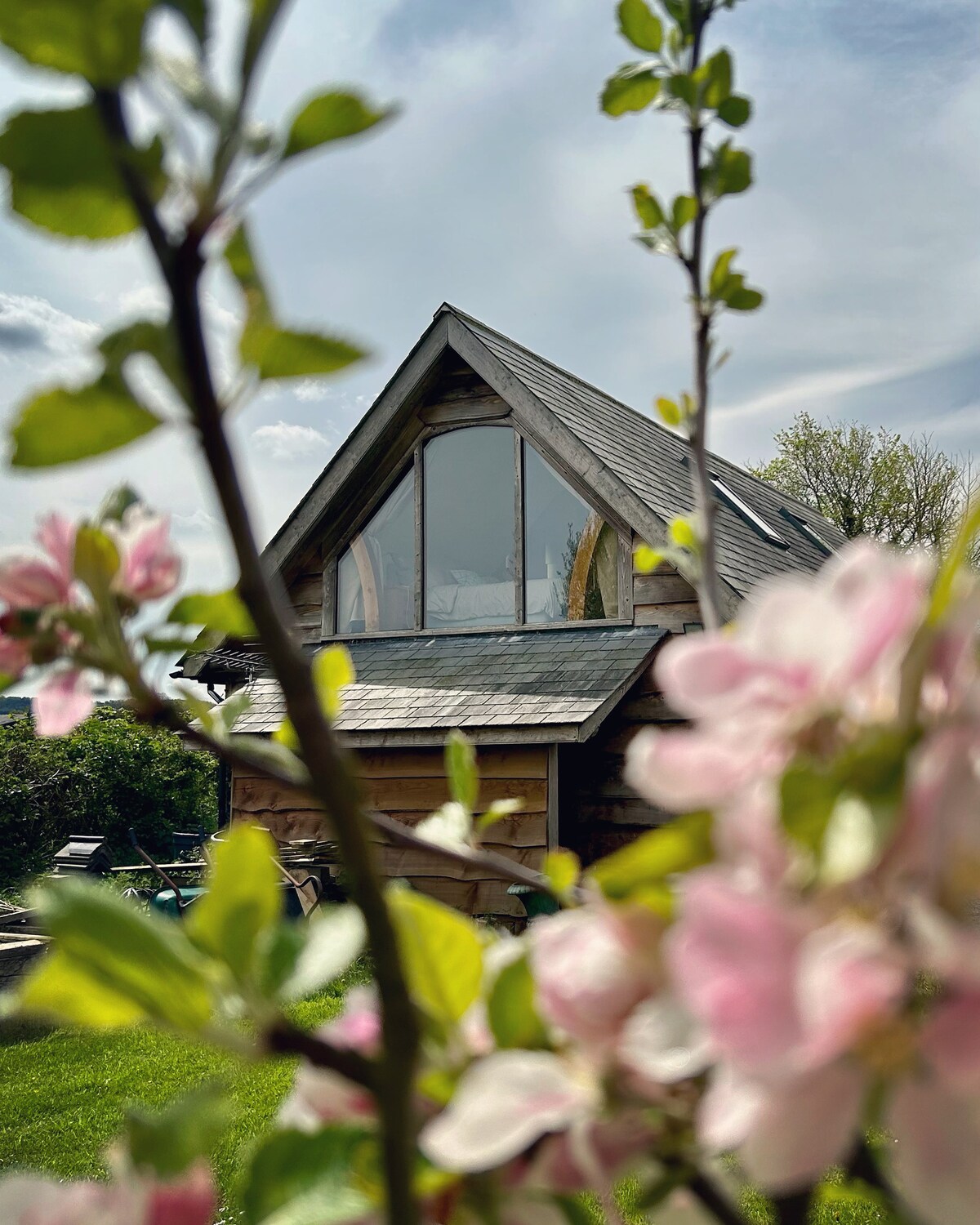 The chalet features a striking gable end window, framed by blossoming branches. Soft pink flowers contrast with the wooden exterior, while lush green grass and hints of outdoor seating are visible in the background, creating a peaceful countryside scene.