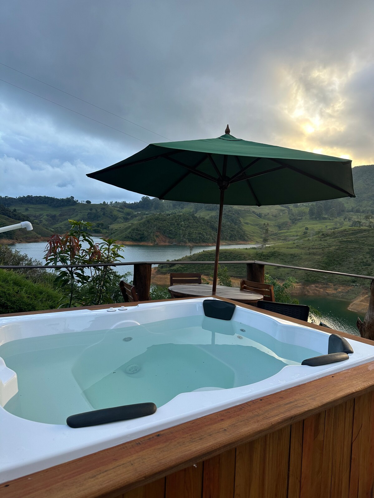 A hot tub is positioned on a wooden deck, accompanied by a green umbrella for shade. The serene landscape of rolling hills and water is visible in the background, with clouds gathering above as the sun sets, creating a peaceful outdoor escape.