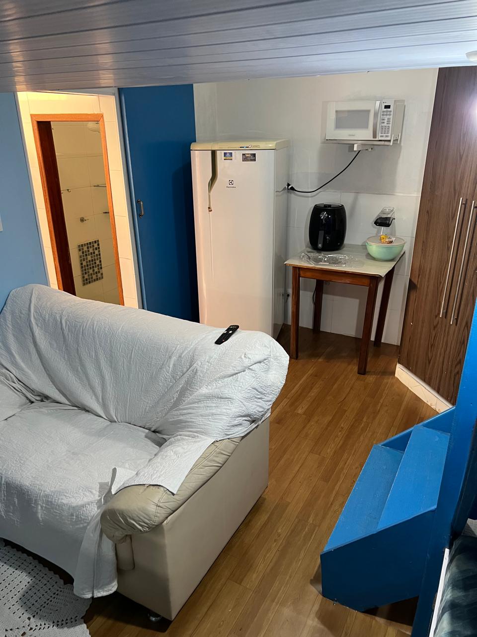A compact kitchen area is visible, featuring a white refrigerator, microwave, and air fryer on a small table. A cream-colored sofa with a white throw is positioned nearby. The light wooden floor adds warmth to the space, with blue walls providing a modern touch.
