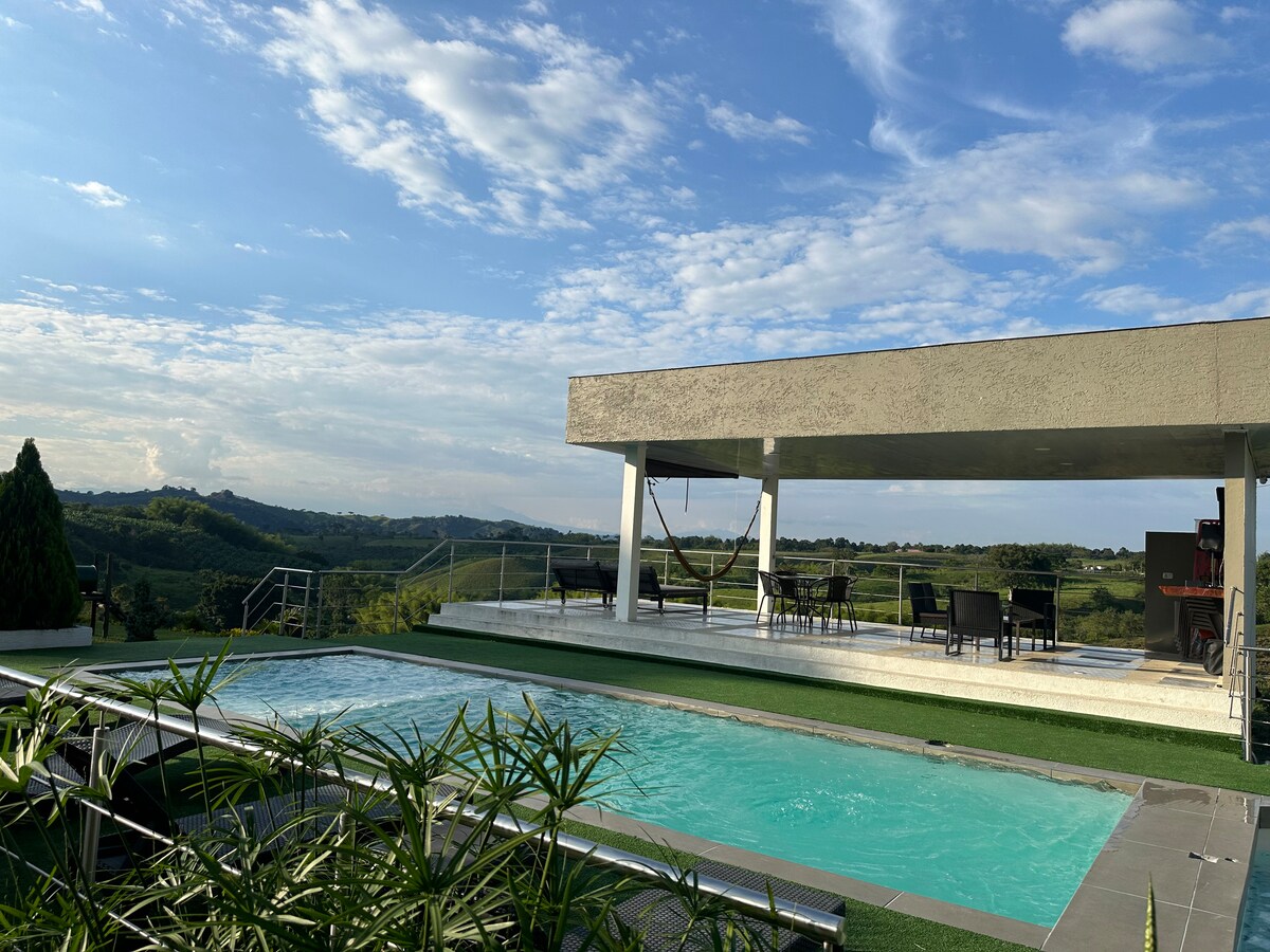 A serene outdoor area showcases a pool surrounded by lush greenery. A covered seating area with chairs is visible, providing a perfect vantage point to appreciate the expansive views of the distant hills under a bright blue sky.