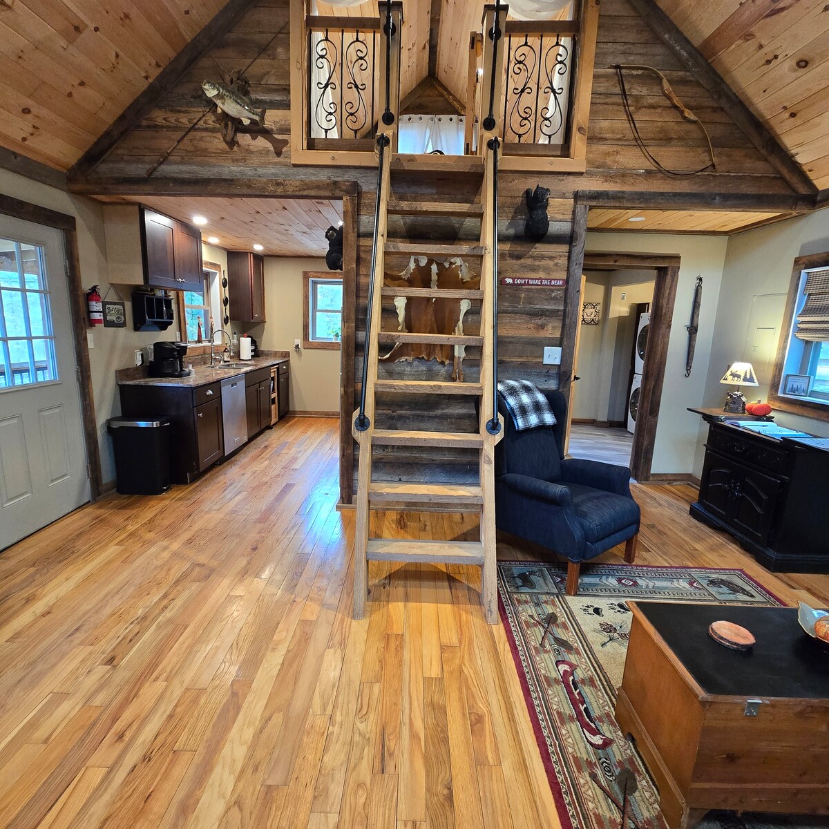 The open layout features a wooden staircase leading to the loft, surrounded by natural wood walls and ceilings. A cozy blue recliner is positioned nearby on an area rug, with kitchen cabinetry visible in the background. Warm wooden flooring enhances the inviting atmosphere.