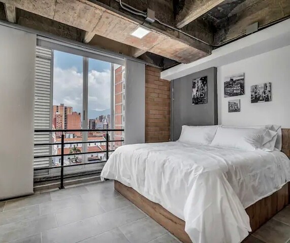 A spacious sleeping area features a large bed with white linens, positioned next to a wide window offering views of the city skyline. Exposed concrete ceilings contribute to an industrial design, while framed artwork adorns the walls.