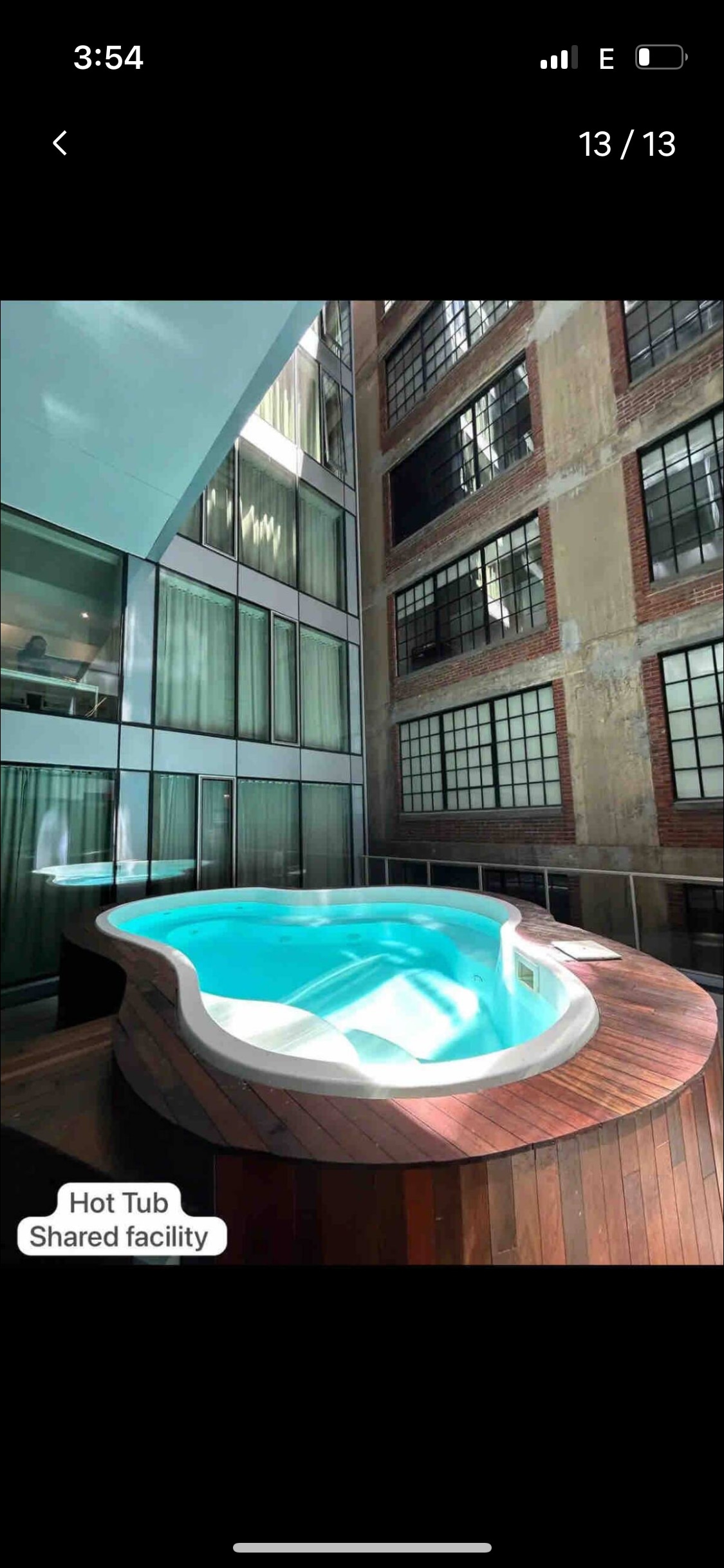 A hot tub occupies a wooden deck, surrounded by tall glass walls that reflect natural light. The space is framed by exposed brick walls, showcasing the building's character. Sheer curtains create a tranquil atmosphere, enhancing the inviting setting for relaxation.