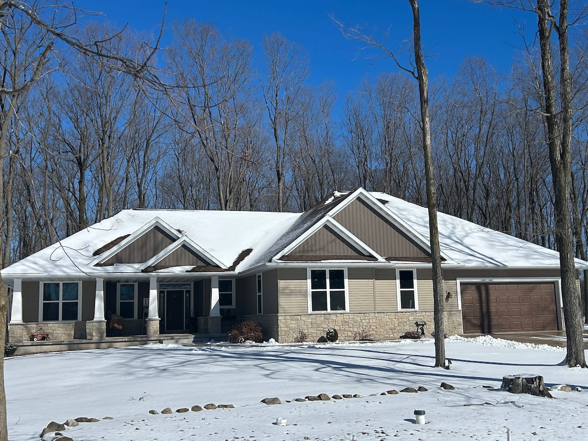 The single-story home is surrounded by bare trees, emphasizing a serene winter setting. A snow-covered roof contrasts against the clear blue sky. The front porch is visible, providing an inviting entrance, and a driveway leads to a single-car garage on the right.