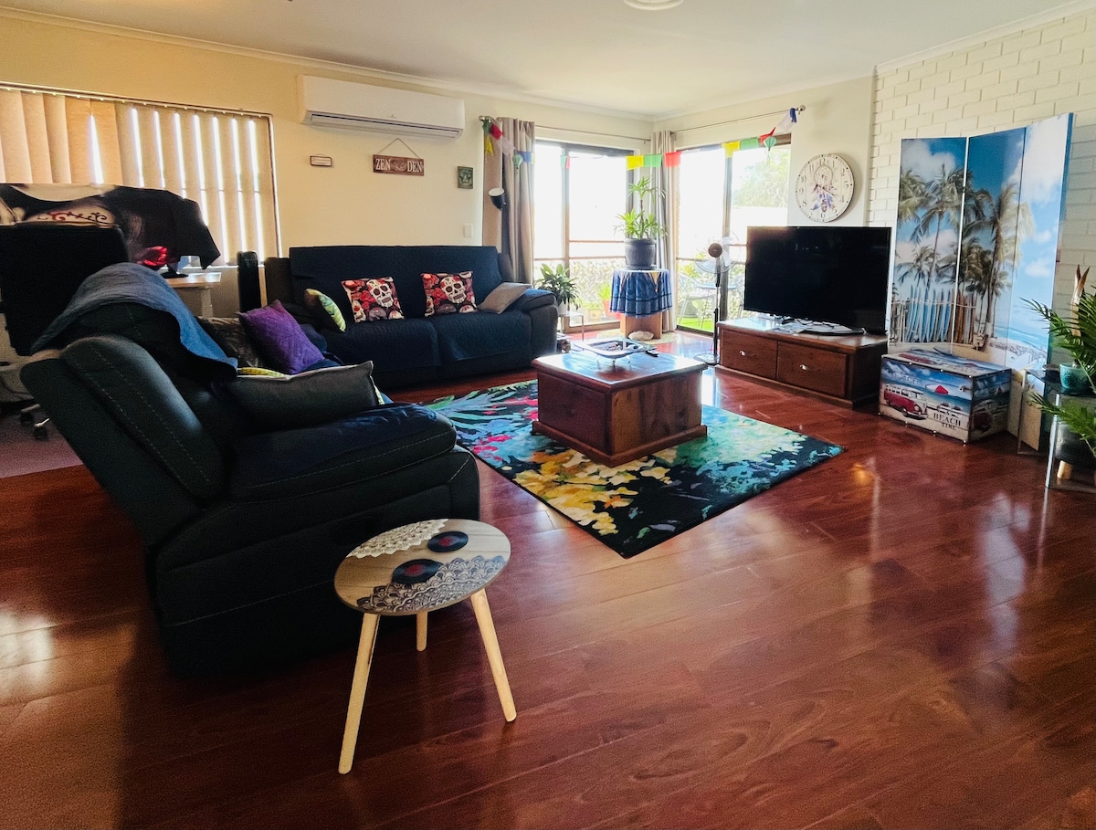 The living area features a dark sofa and armchair, with a colorful area rug beneath a wooden coffee table. Light is present from large windows, showcasing outside views. Decorative accents include wall art and plants, enhancing the relaxed ambiance of the space.