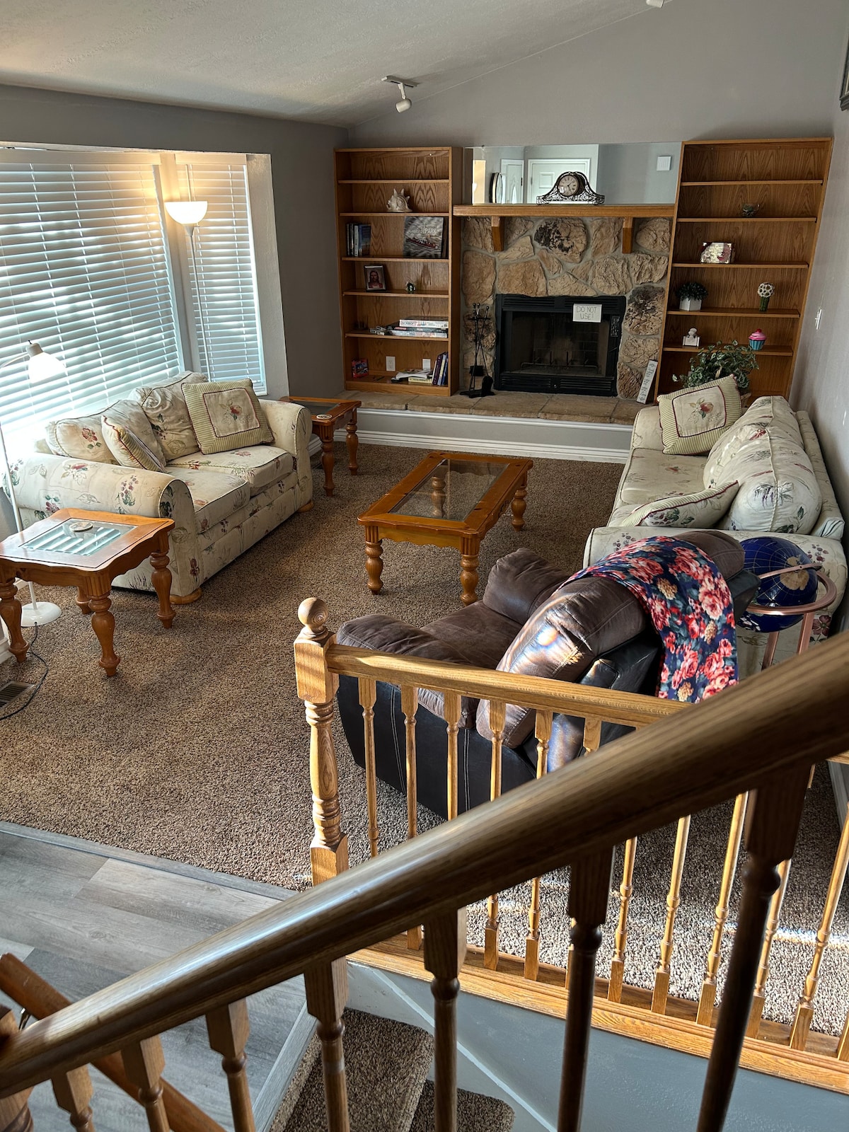 A cozy living room is showcased with two floral patterned sofas and a dark leather recliner. A wooden coffee table is centered on a plush area rug. The space features built-in shelves and a fireplace, while natural light streams through large windows.