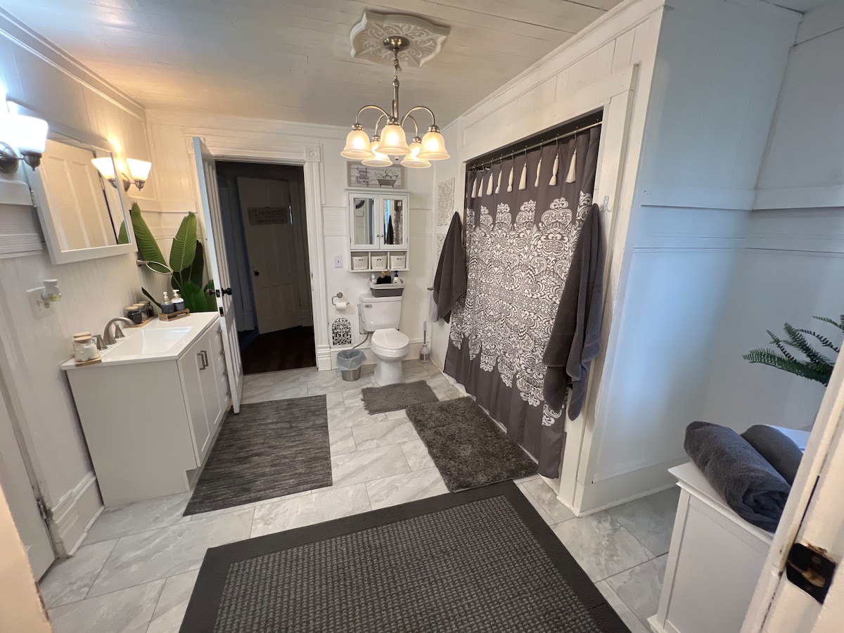 A spacious bathroom features marble flooring and a neutral color palette. A large mirror is mounted above a vanity, and a decorative shower curtain hangs beside neatly arranged towels. Two light fixtures provide ample illumination, and plants add a natural touch to the decor.