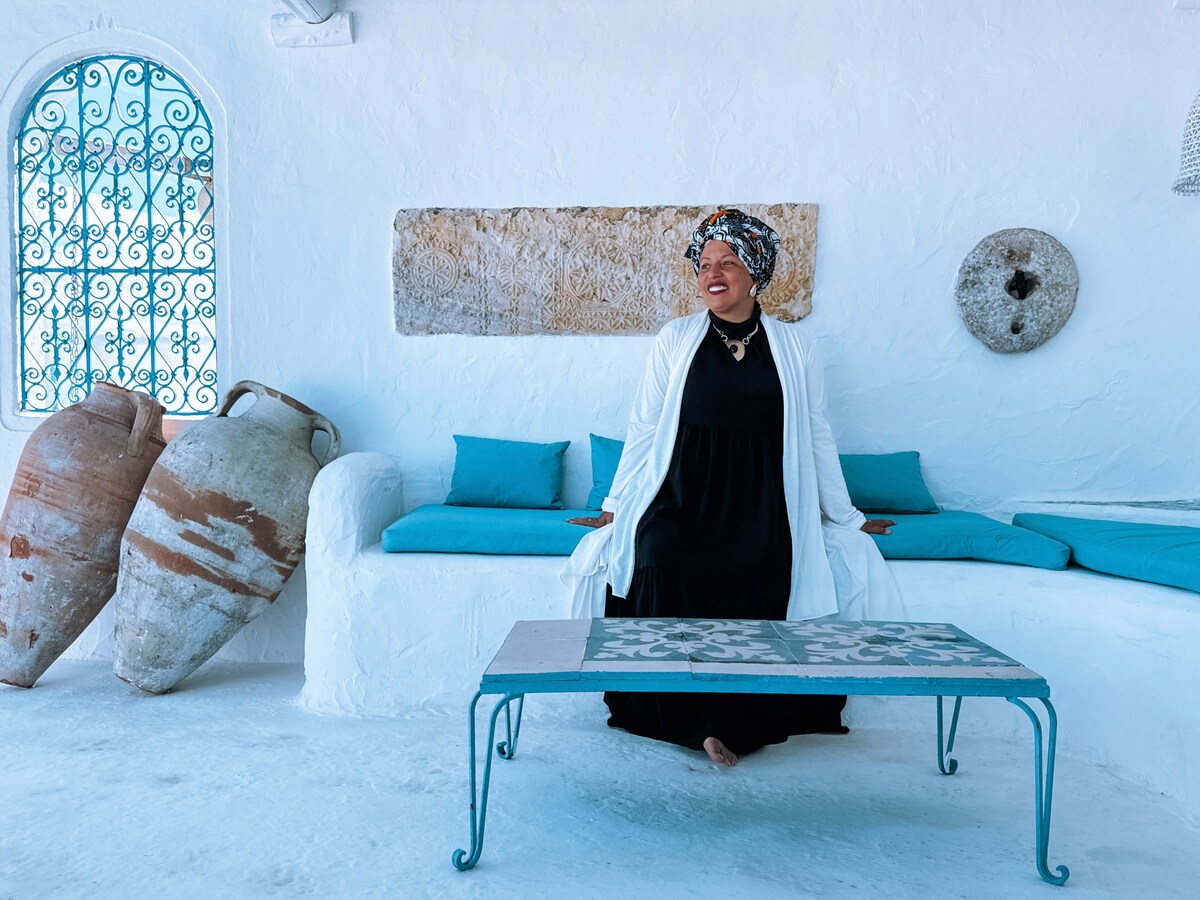 A serene seating area is presented with whitewashed walls and contrasting colorful cushions. A decorative table stands in the center, surrounded by traditional pottery, while an intricate window frame allows soft light to filter through.