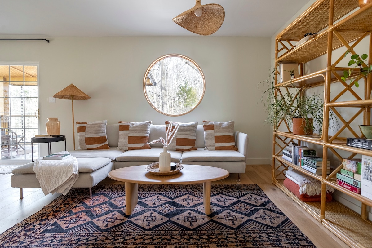 A spacious living area is characterized by a light-colored sectional sofa adorned with patterned cushions. An oval window allows natural light to fill the room, while a round coffee table sits on a patterned area rug. A decorative shelf displays books and plants, adding to the inviting ambience.