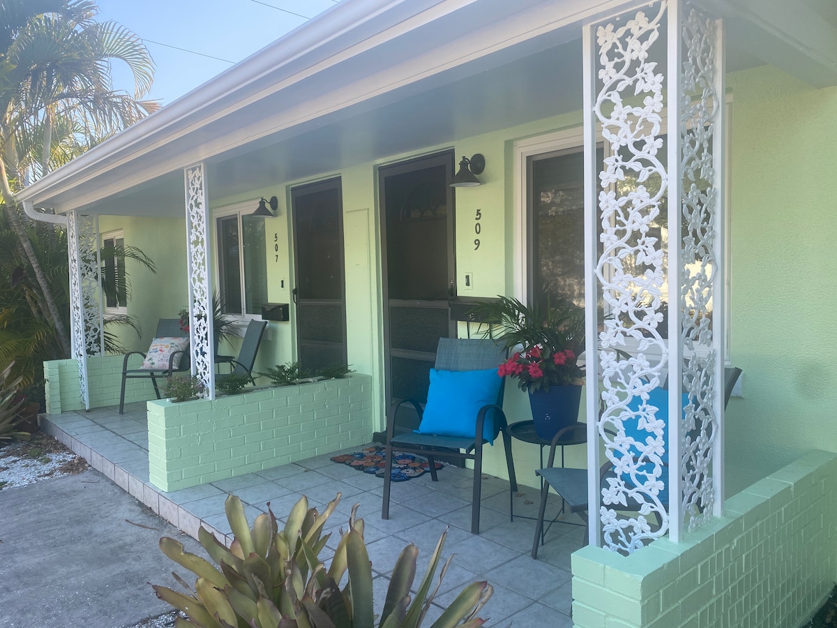 A welcoming porch space is depicted, featuring pastel green walls and decorative metalwork. Two chairs and a small table are positioned on the tiles, with potted plants adding color to the environment. The entrance is framed by a sense of openness, inviting relaxation.