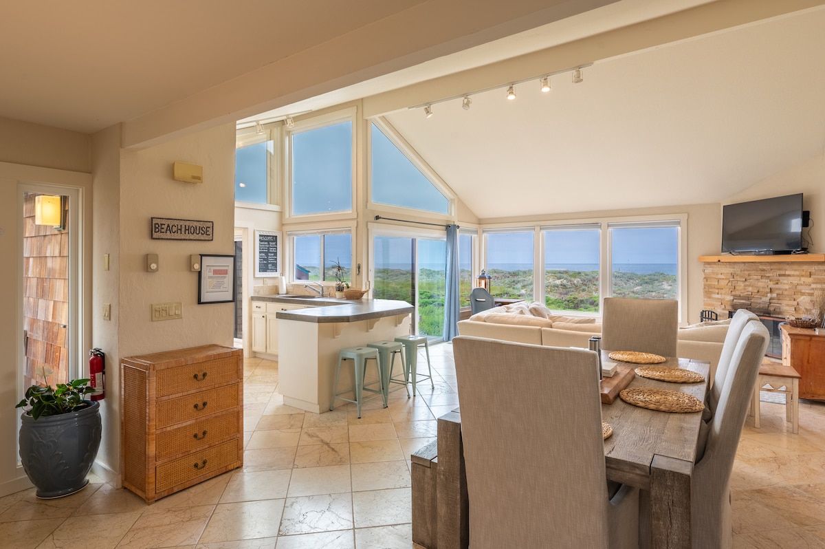 A bright and airy living space features high vaulted ceilings and panoramic windows that showcase views of the Pacific Ocean. Light-colored tiles enhance the open layout, which includes a dining area and kitchen with modern bar seating. Natural light floods the room, creating a welcoming ambiance.