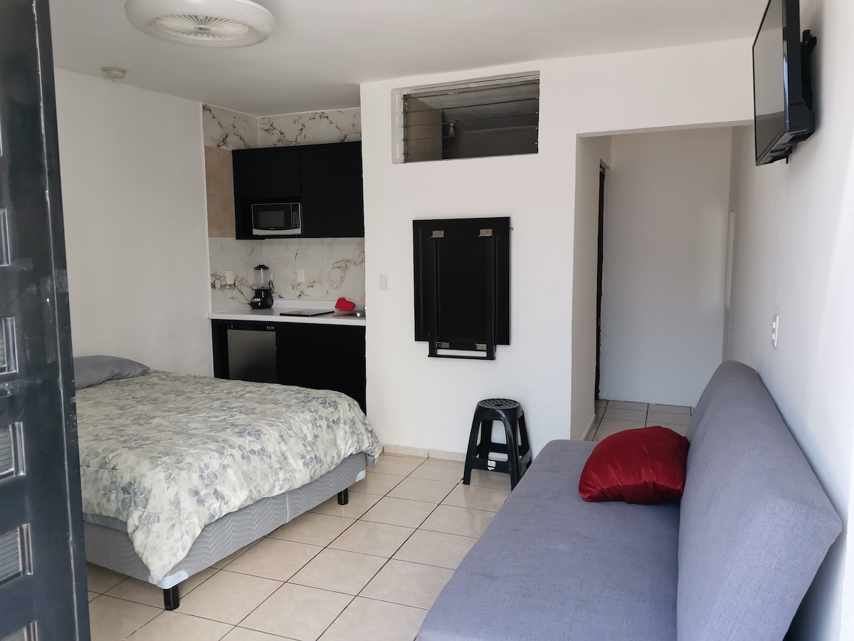 A simple and functional living space features a spacious bed with a floral quilt. An adjacent kitchenette includes essential appliances. A gray sofa sits against the wall, providing additional seating. Natural light enters through the window, creating a bright and open atmosphere.