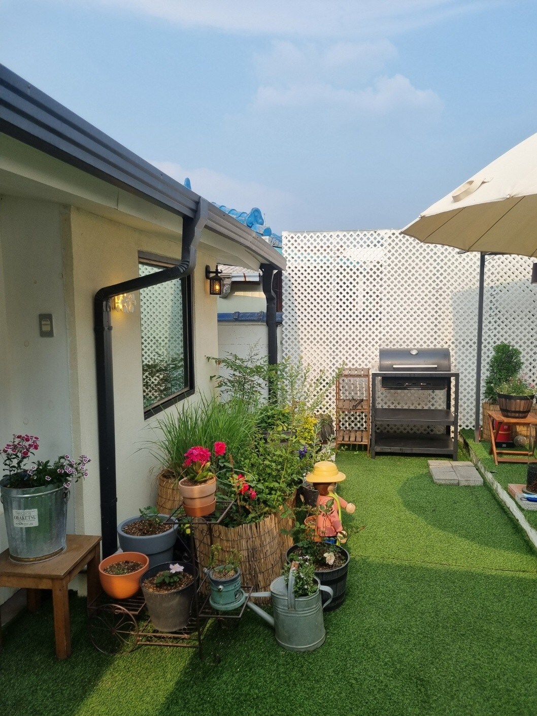 A charming garden area is depicted, featuring lush greenery and colorful flower pots arranged along the edges. A patio umbrella provides shade over a small seating area. A barbecue grill is visible, complemented by neatly organized outdoor decor.