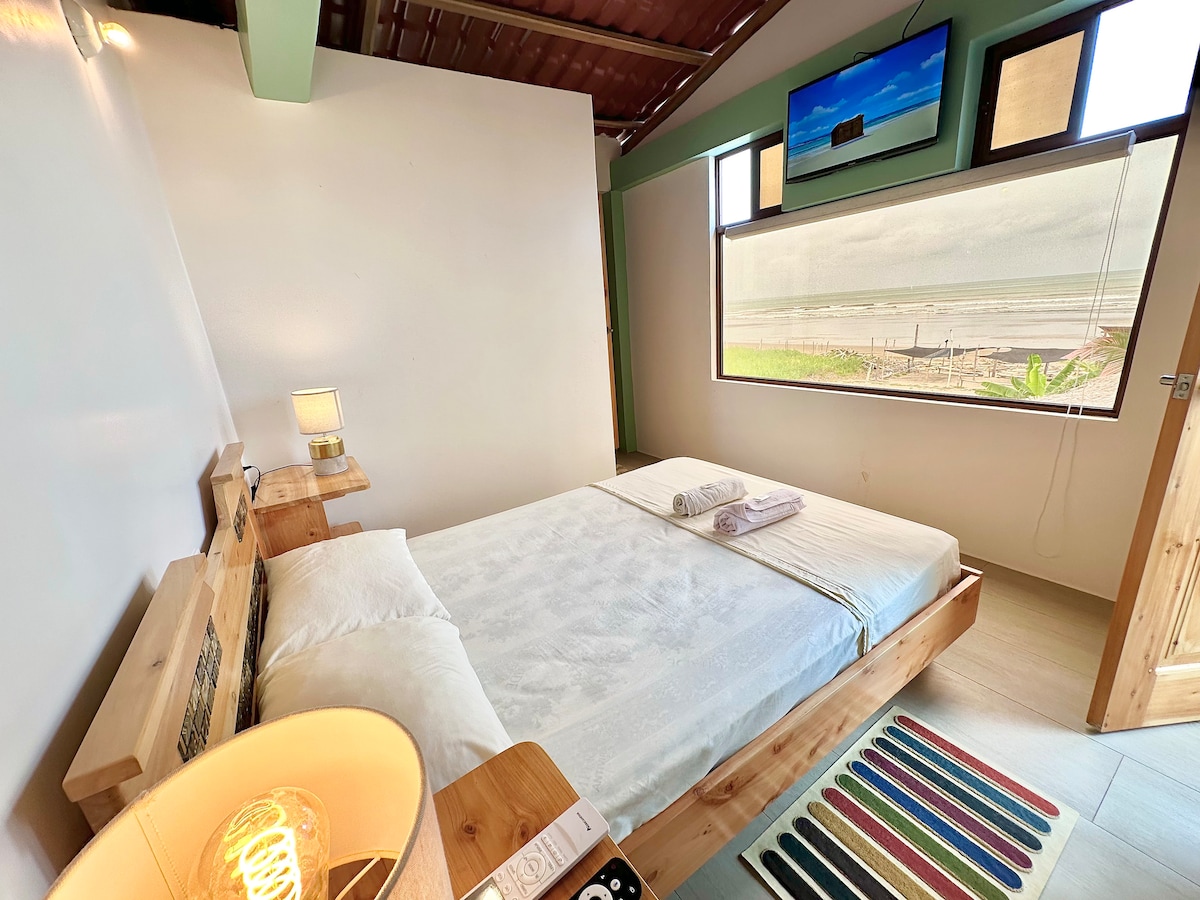 A comfortable bedroom is presented with a spacious bed featuring soft linens. A large window offers a view of the beach and ocean, while a small bedside lamp and decorative rug add warmth to the space. A television is mounted on the wall.