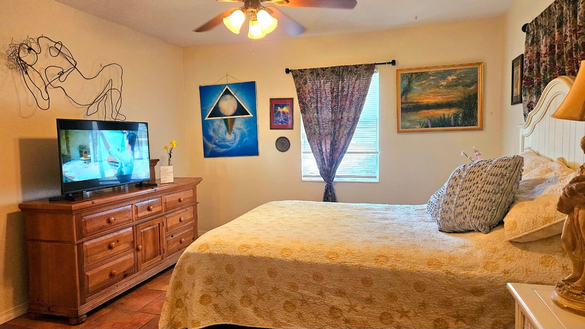 A private bedroom features a queen-sized bed with a light quilt, complemented by decorative pillows. A dresser supports a TV, while artwork adorns the walls. Soft natural light enters through a window covered by sheer curtains, enhancing the serene atmosphere.