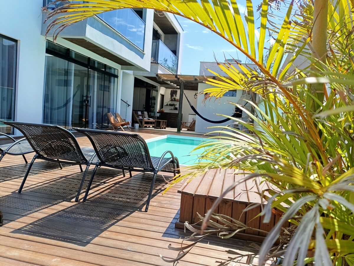 A serene pool area is surrounded by wooden decking, featuring several lounge chairs positioned for relaxation. Lush greenery frames the scene, with the clear water of the pool invitingly reflecting sunlight. The backdrop includes a modern house with expansive glass doors.