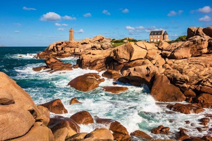 A coastal scene features rugged, large rocks with crashing waves below. A lighthouse stands prominently in the background, next to a charming stone house. The sky is bright with scattered clouds, conveying a serene maritime atmosphere.