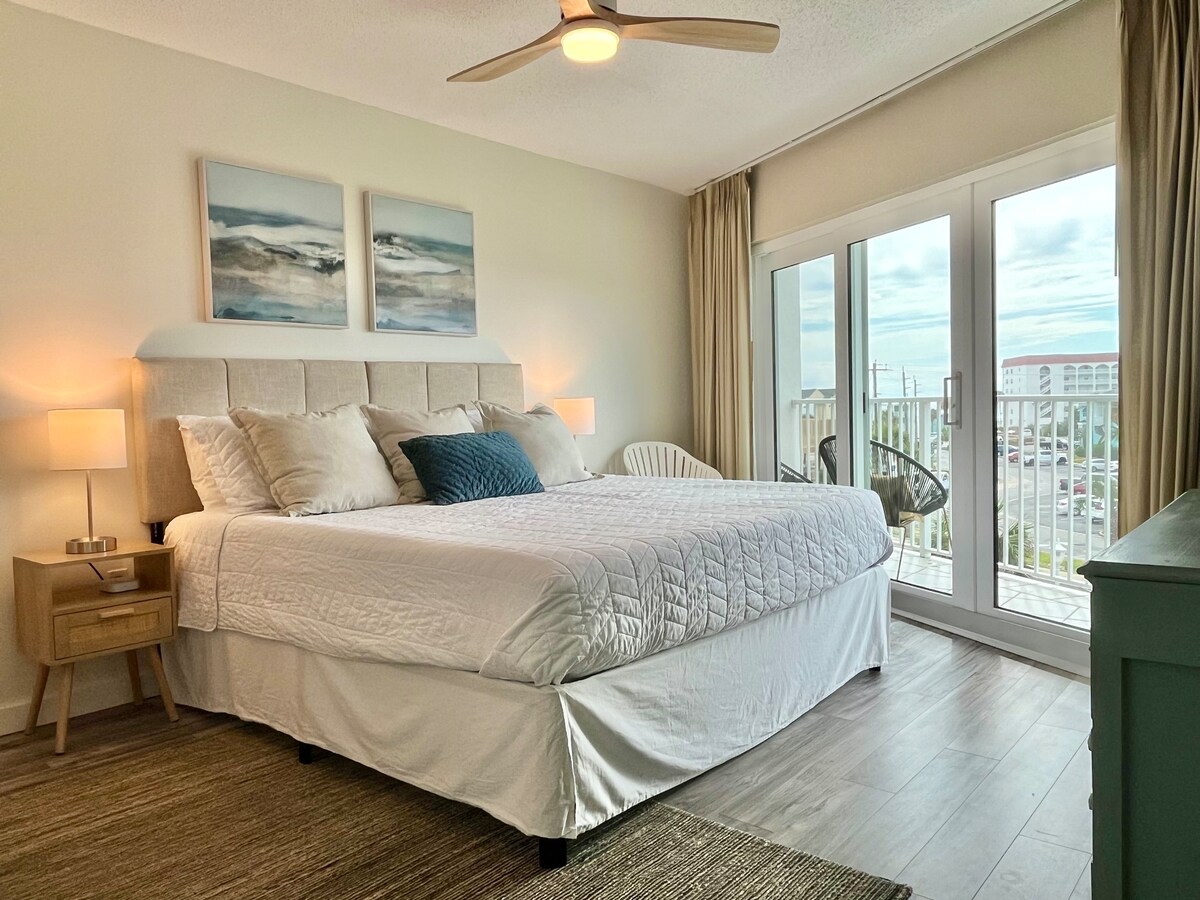 A spacious bedroom features a king-size bed dressed in soft linens, accompanied by two bedside lamps. Large glass doors provide access to a balcony, allowing natural light to brighten the room. A comfortable chair is positioned nearby, enhancing the inviting atmosphere.
