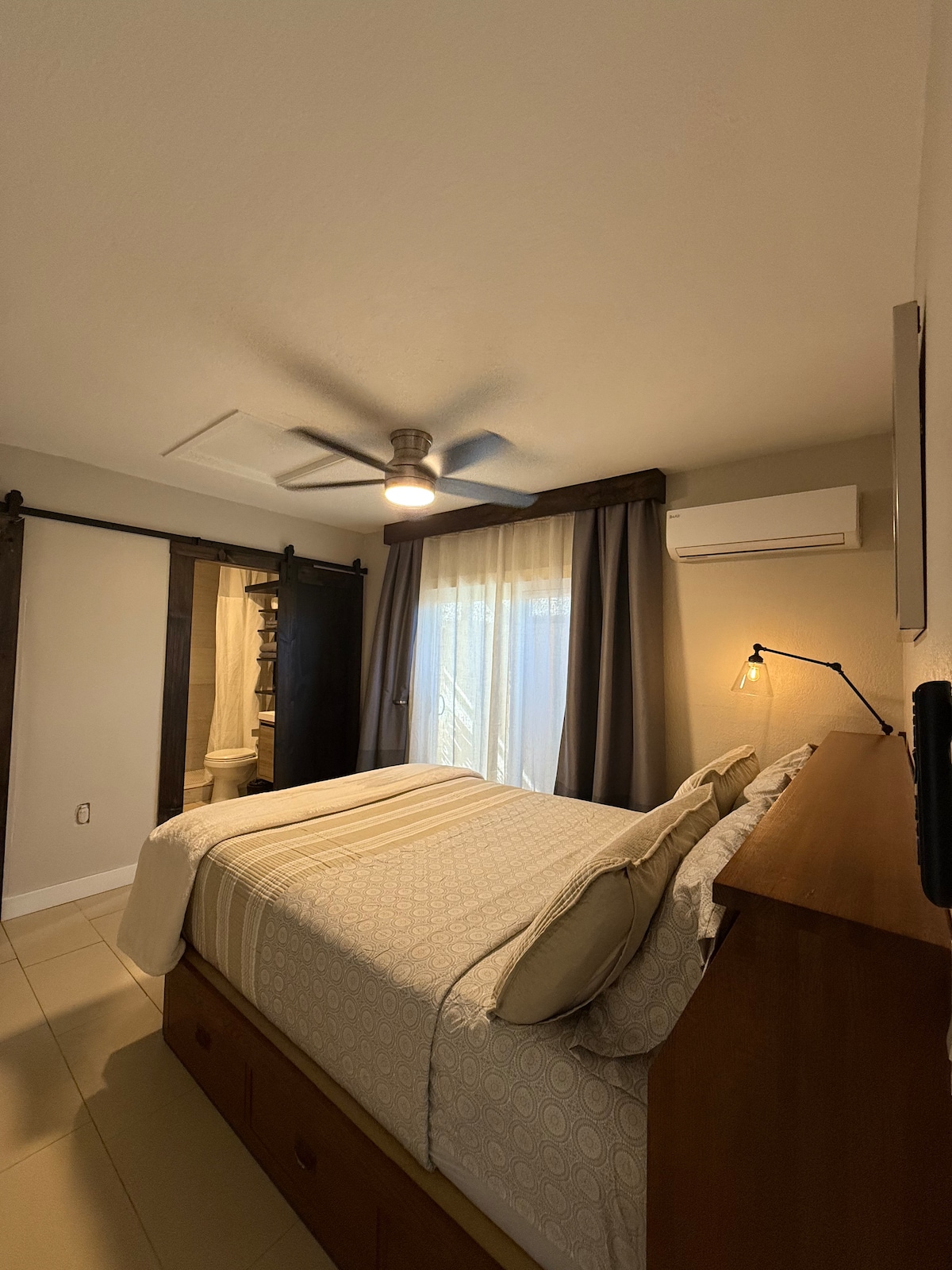 A well-lit bedroom features a spacious bed with decorative pillows and a soothing quilt. A ceiling fan hangs above, and natural light filters through sheer curtains. A sliding door leads to a bathroom area, complementing the room's modern design.
