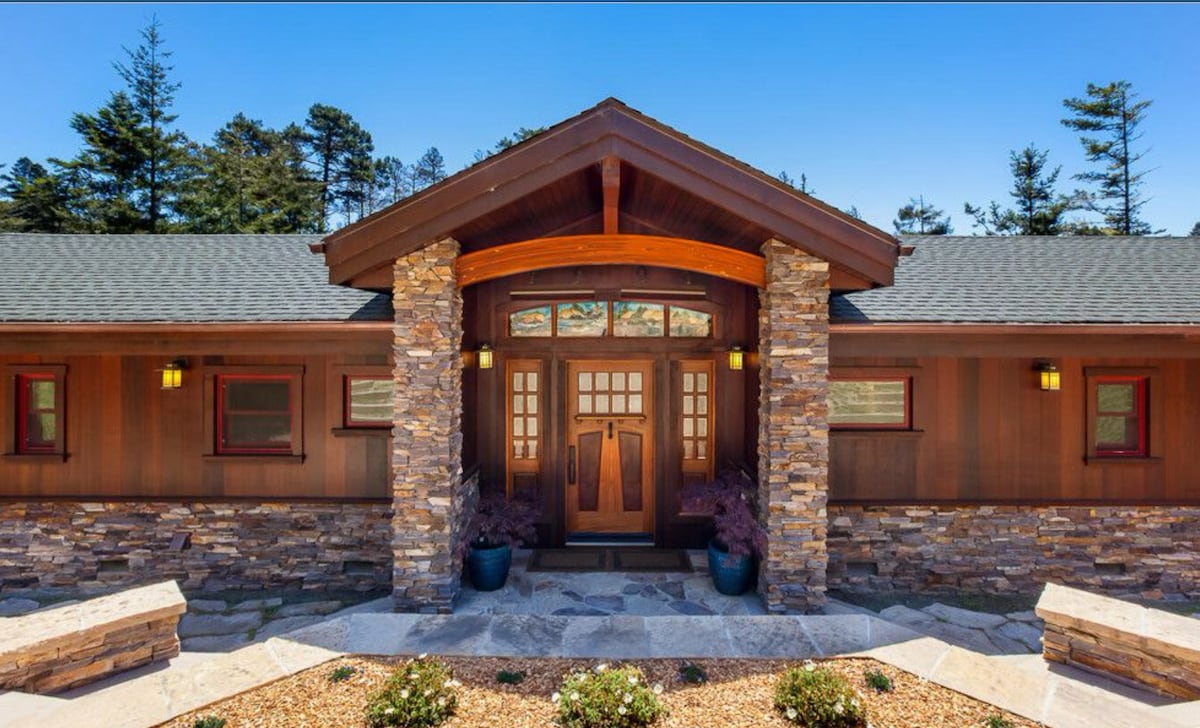 The entrance features a wooden structure with stone accents, framed by lush greenery. The double doors are adorned with glass panels, and pendant lighting enhances the welcoming atmosphere. Stone pathways lead towards the front entrance, surrounded by landscaped garden beds with decorative plants.