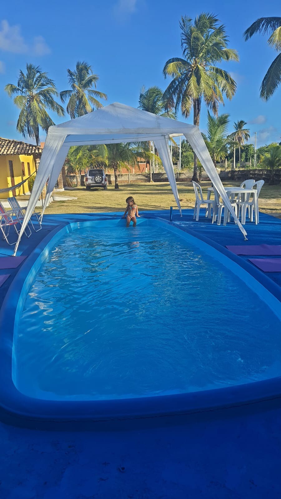 A clear swimming pool is featured under a spacious canopy, surrounded by a well-maintained garden with palm trees. Poolside chairs are positioned thoughtfully on the patio. Sunlight reflects off the water, creating a serene ambiance, inviting relaxation and enjoyment.