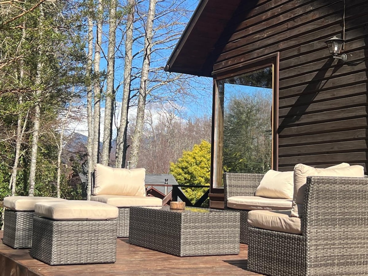 An outdoor seating area features comfortable wicker furniture with cream cushions. A coffee table sits in the center, surrounded by lush greenery and trees, with a cabin's wooden facade visible in the background under a clear blue sky.