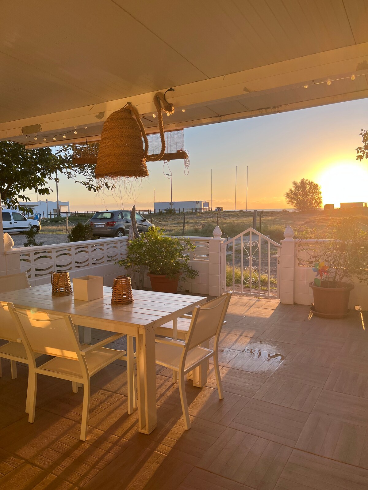 A patio area features a wooden dining table surrounded by white chairs, adorned with lanterns. Sunlight casts a warm glow as the sun sets in the background, illuminating the landscape beyond the white gate and lush greenery.