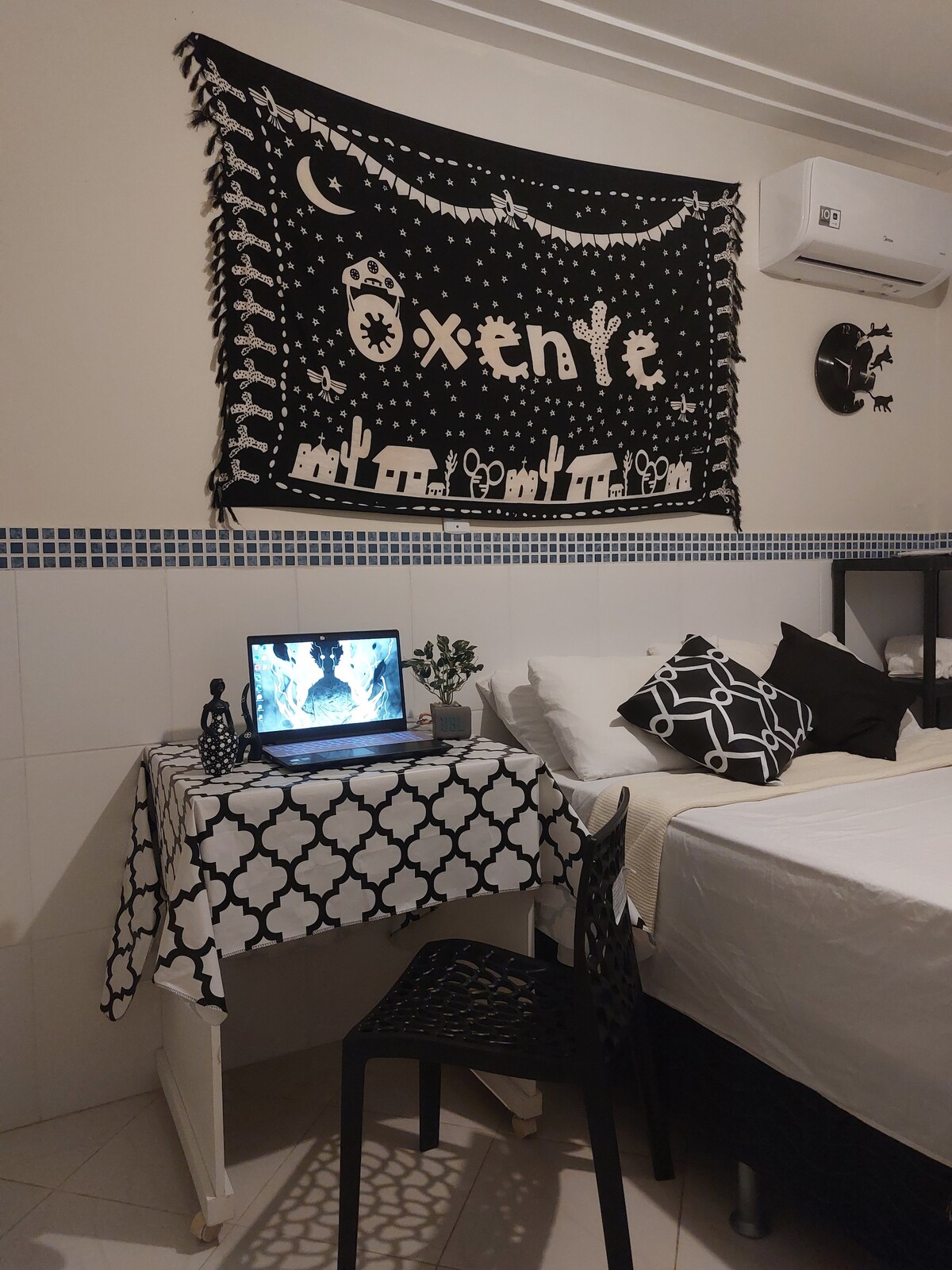 A well-furnished room includes a bed alongside a small desk with a laptop. A decorative wall hanging featuring a desert scene is placed above the desk. A black chair and a small plant contribute to the room's simple design. Air conditioning is installed on the wall.