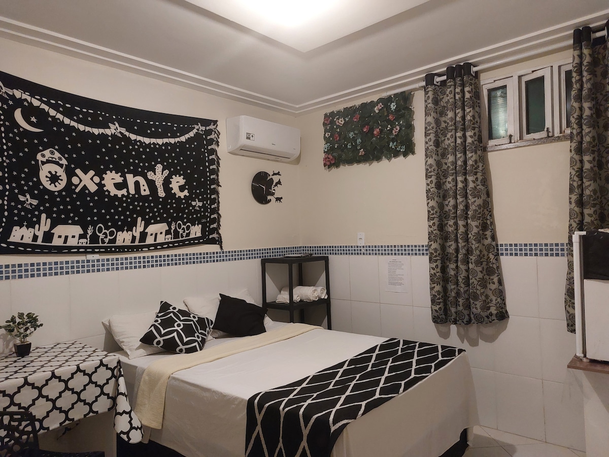 A well-furnished room features a spacious bed with black and white bedding. A small table with a plant is positioned next to the bed. Dark curtains frame a window, while a wall art piece adds a decorative touch. Air conditioning is present for comfort.