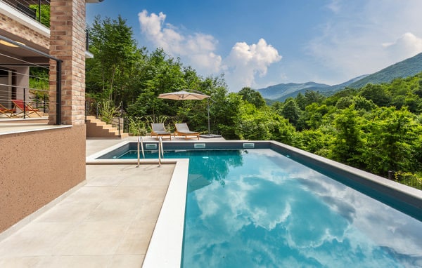 An outdoor swimming pool is framed by a spacious deck, offering sun loungers and an umbrella for shade. Lush greenery surrounds the area, while distant mountains provide a scenic backdrop. The clear blue water reflects the sky, enhancing the serene atmosphere.