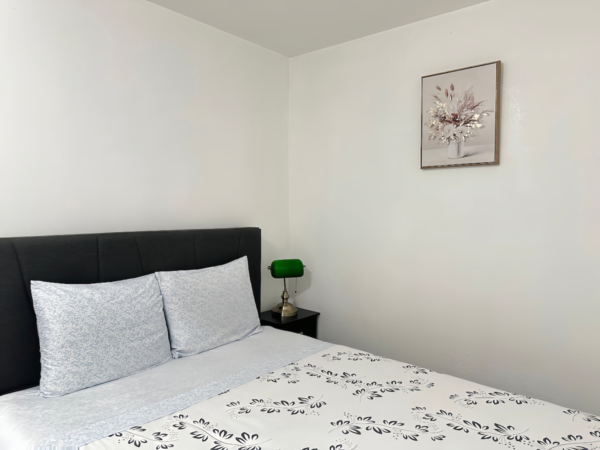 A private bedroom is displayed featuring a queen-size bed with clean bedding and decorative pillows. A nightstand with a lamp and a framed flower artwork adorns the white wall, enhancing the room's overall simplicity and tranquility.