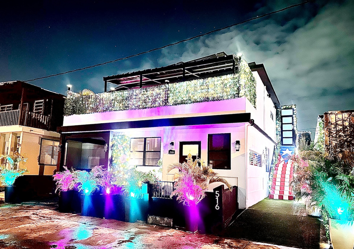 The exterior of the modern casita is illuminated at night, featuring vibrant lights and lush greenery. A spacious terrace is visible on the second floor, while the building showcases clean lines and a welcoming facade, enhancing the overall charm of the neighborhood.