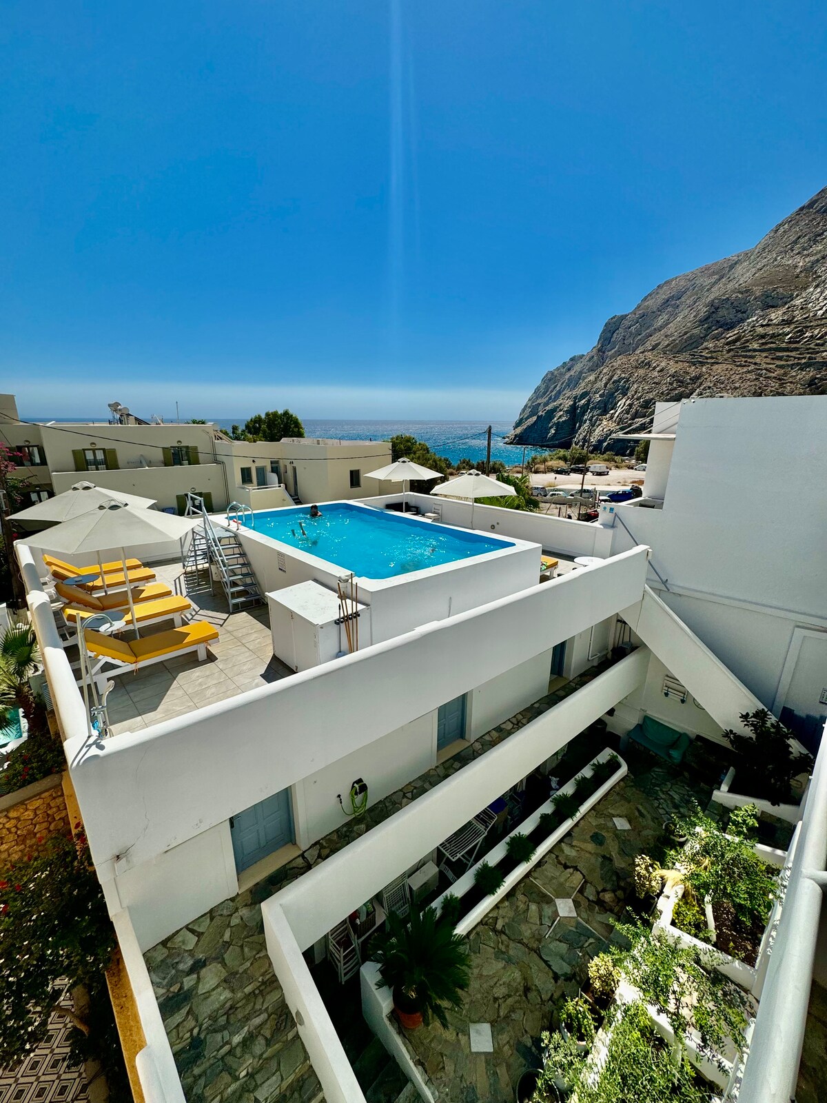 An aerial view captures a shared swimming pool surrounded by sun loungers. The ocean glimmers in the distance, with mountain silhouettes framing the scene. White-walled buildings and greenery enhance the tranquil outdoor space.