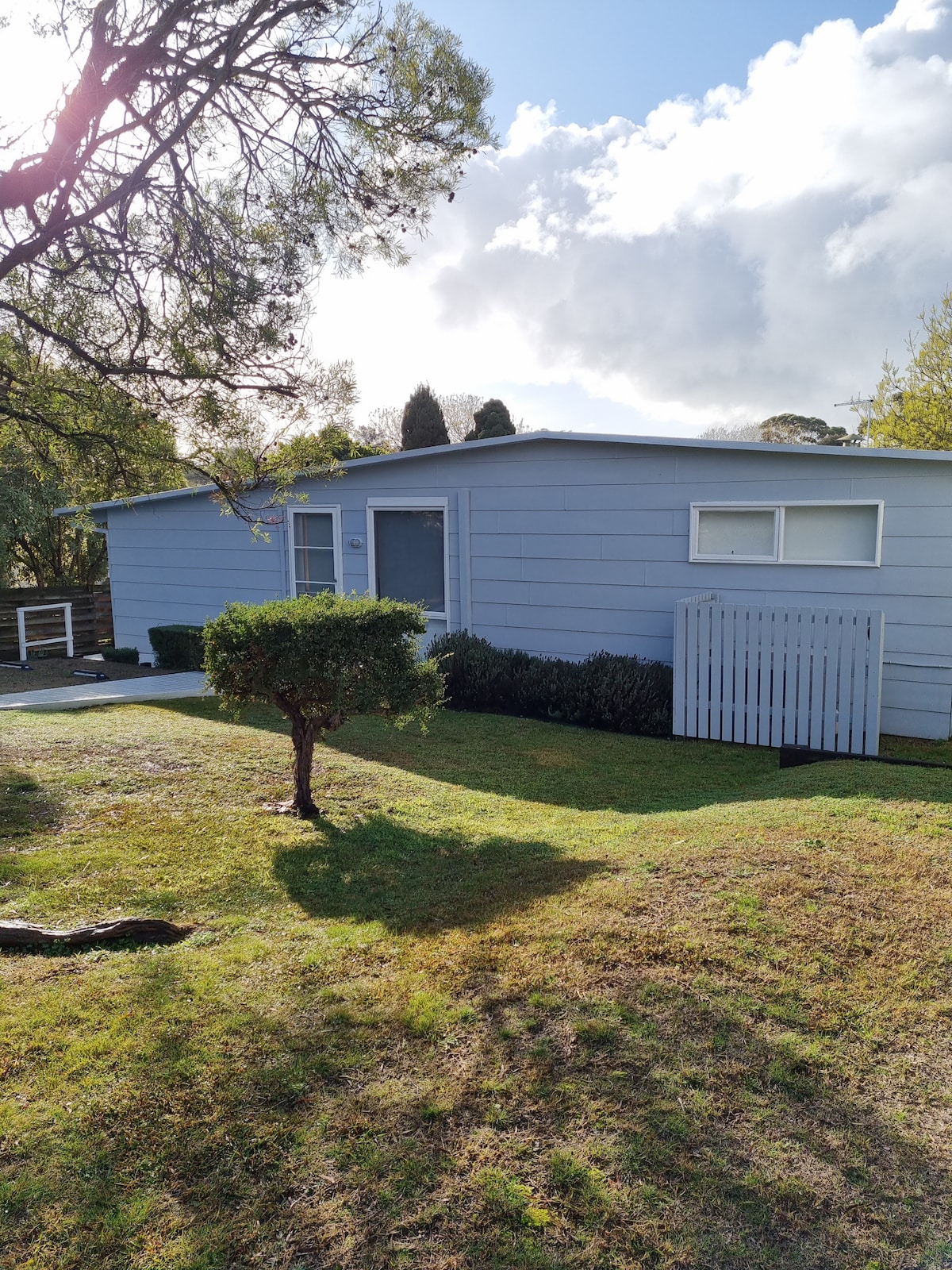 A light blue beach house is set amidst grassy landscaping, featuring a well-kept front yard with shrubs and small trees. The sunny exterior is complemented by clouds in the background, providing an inviting appearance. A white picket fence adds charm to the property.