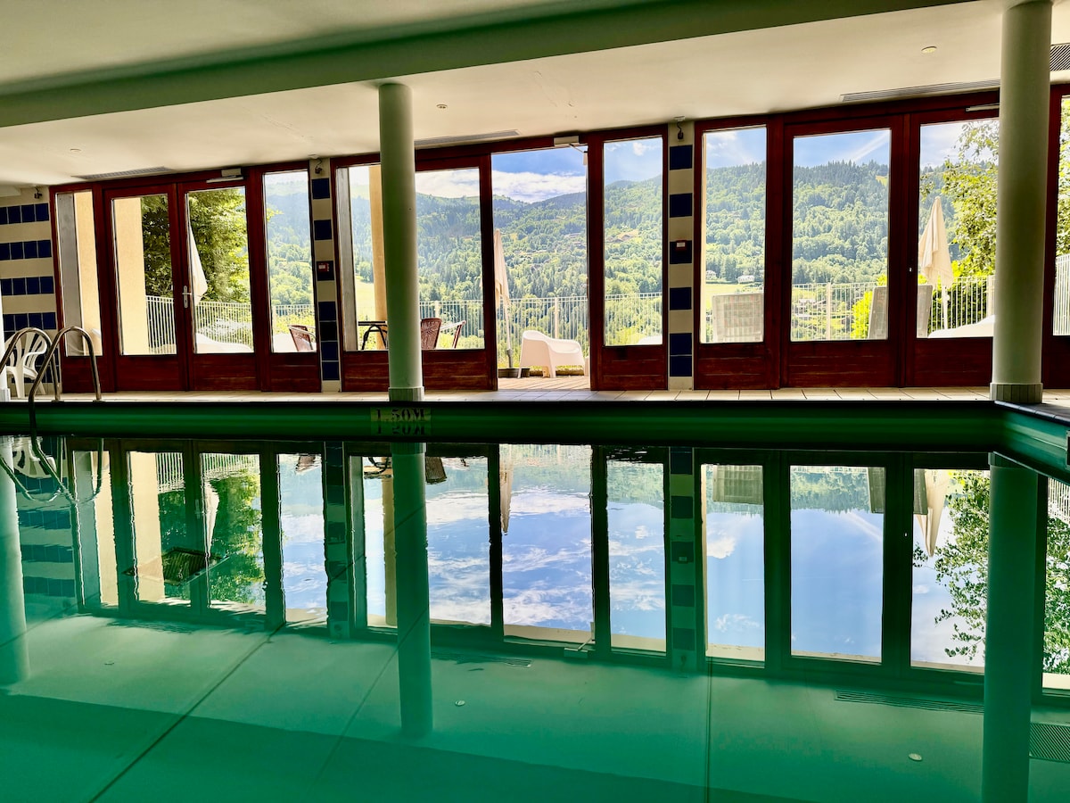 An indoor heated pool reflects the surrounding landscape through large glass windows. Comfortable seating is visible outside on a terrace, offering a view of the green hills and trees. The setting creates a serene atmosphere for relaxation and leisure.