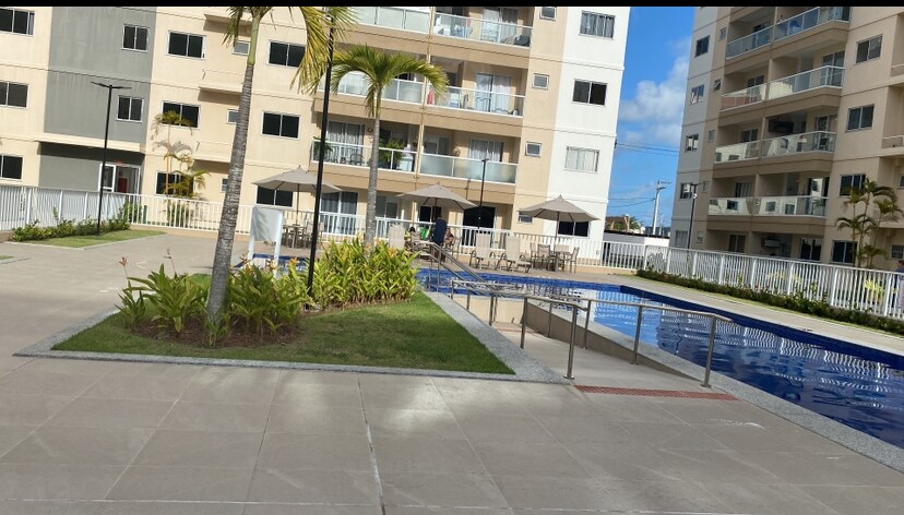 A well-maintained outdoor area features a sleek swimming pool surrounded by manicured gardens. Lounging areas with umbrellas and tables are visible, alongside a pathway leading towards the apartments. Palm trees and greenery enhance the serene atmosphere of the space.