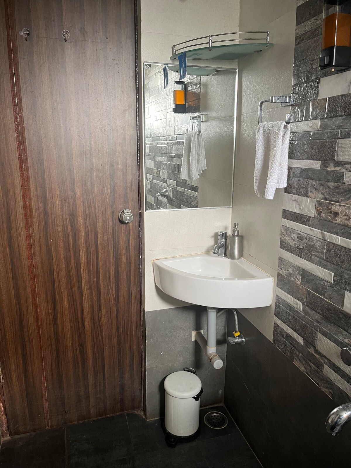 A bathroom features a modern sink with a wall-mounted mirror above it. The wall is adorned with textured stone tiles, and a wooden door provides privacy. A small waste bin and hand towels are visible, enhancing functionality and convenience.