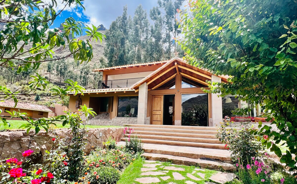 A charming house features a welcoming entrance with a large wooden door and a covered porch. Stone steps lead to the front door, flanked by blooming flowers and lush greenery. Towering trees and scenic hills are visible in the background, enhancing the natural setting.
