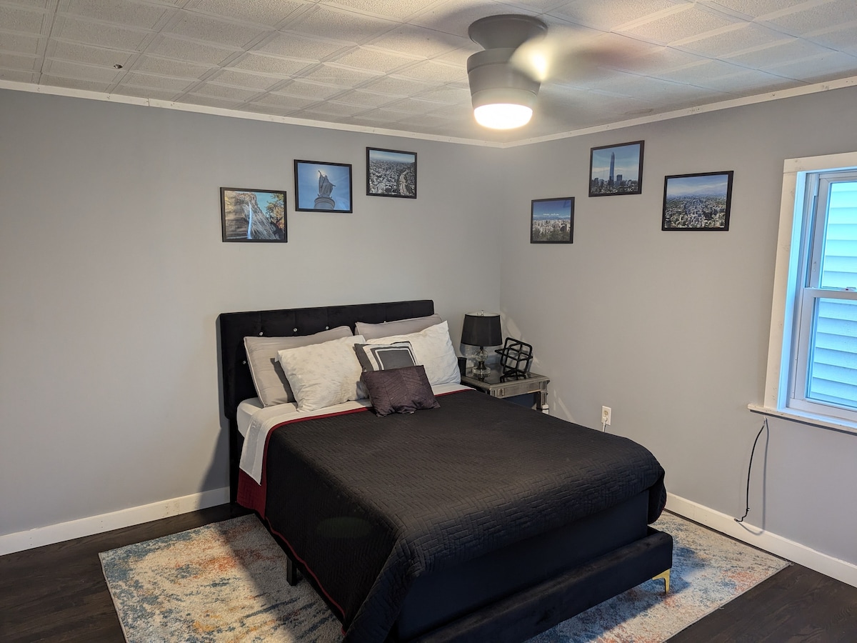 A spacious bedroom is accented with a dark bedspread over a plush mattress. Framed cityscape photographs adorn the light gray walls. A nightstand holds a lamp, and a window on the side allows natural light to fill the room. Wood flooring is visible beneath an area rug.