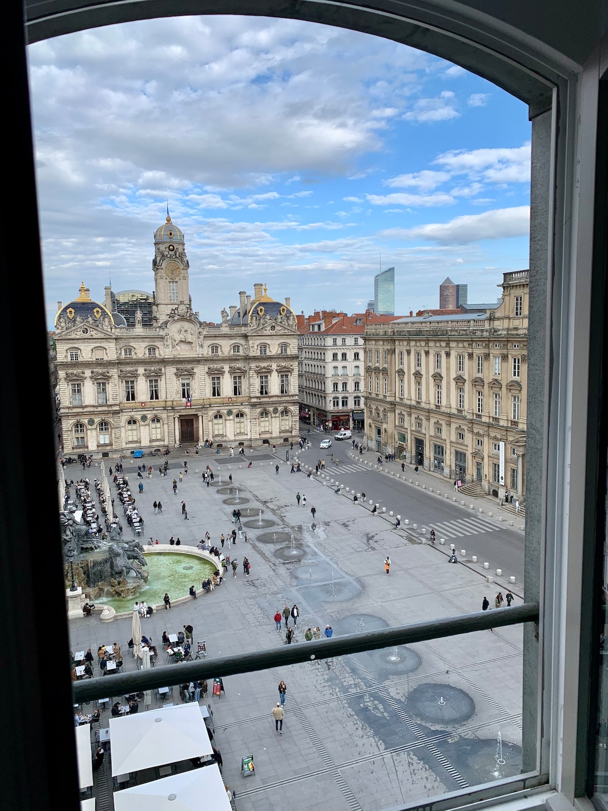 A view is captured from a window, showcasing a vibrant city square. Historic buildings frame the scene, with a fountain at the forefront. People are seen enjoying the open space, while a mix of architectural styles is visible in the background against a clear blue sky.