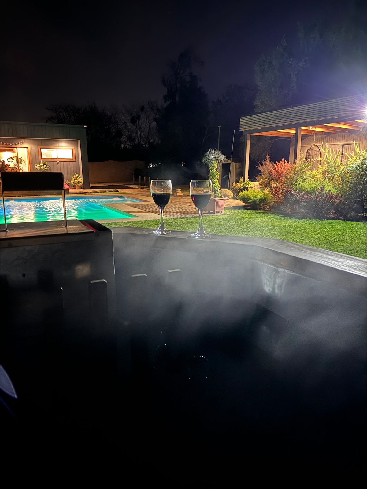 Two glasses are placed near the edge of a hot tub, surrounded by a misty atmosphere. In the background, the illuminated pool and outdoor seating area are visible, showcasing a lush, green landscape under subtle nighttime lighting.
