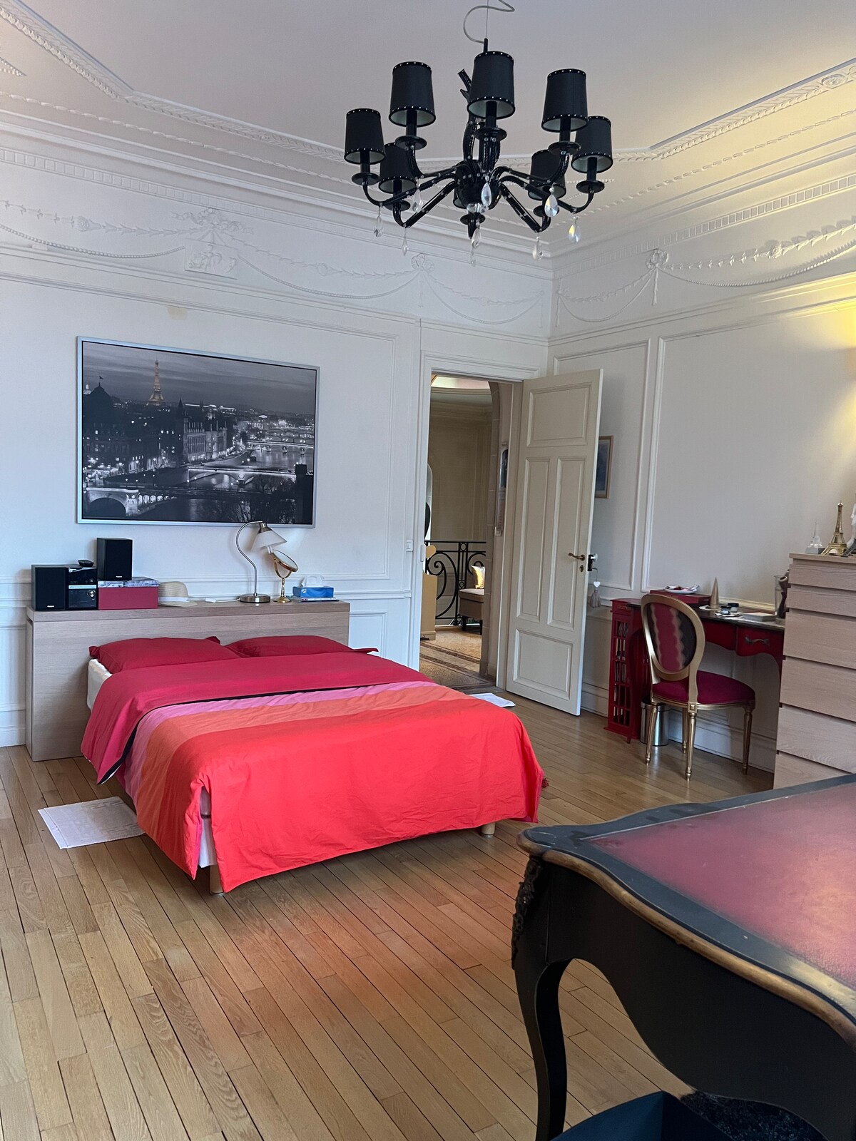 A spacious sleeping area features a vibrant red bedspread over a large bed. Elegant wood flooring is visible, complemented by a stylish chandelier overhead. A dresser and desk with a chair are neatly arranged, while a black and white photograph adorns the wall.