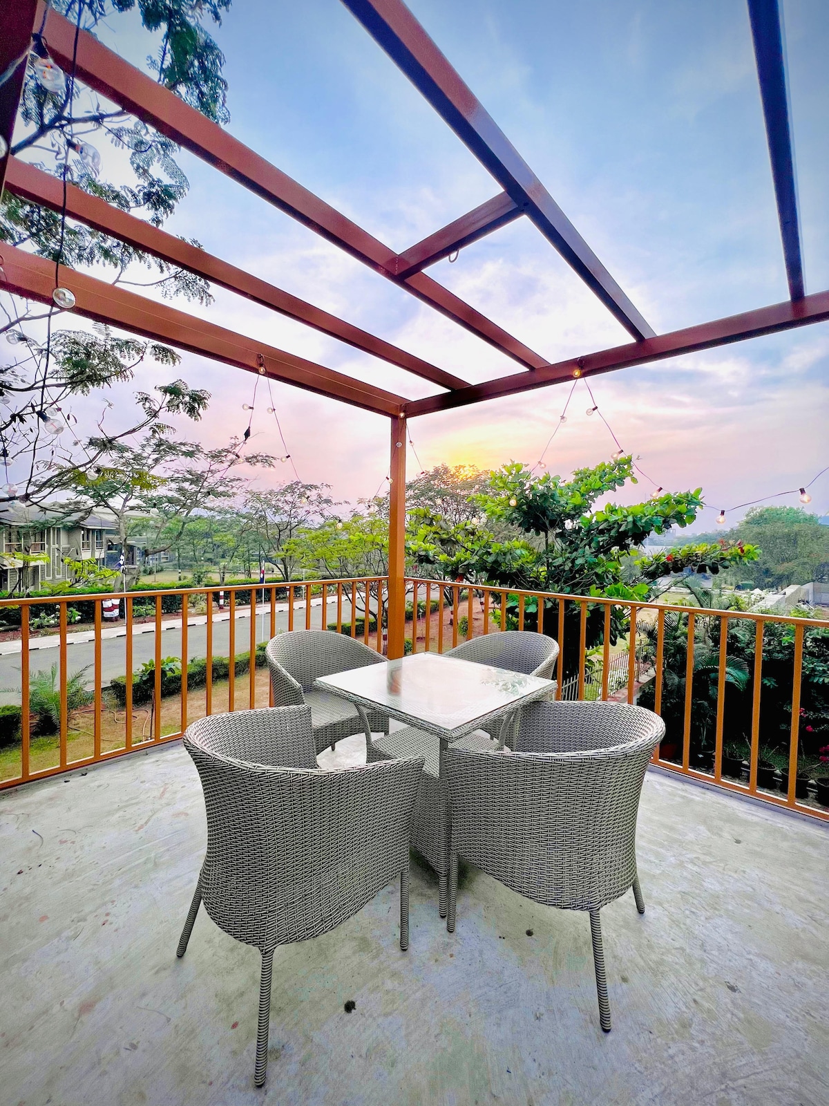 A covered outdoor terrace features a glass-top dining table surrounded by four woven chairs. The area is adorned with string lights, and green foliage is visible in the background, with a soft sunrise illuminating the sky.