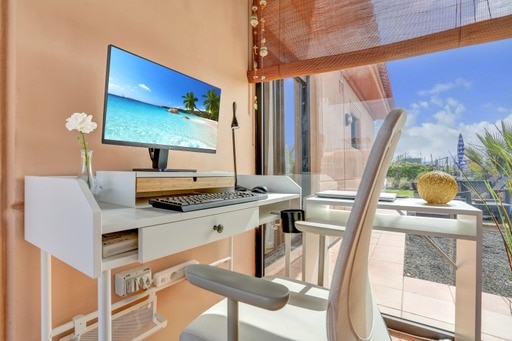 A bright workspace is featured with a desk facing a window, displaying a computer monitor with a beach scene. A potted flower adds a touch of nature, while the surroundings include a view of palm trees and blue skies, creating a serene working environment.