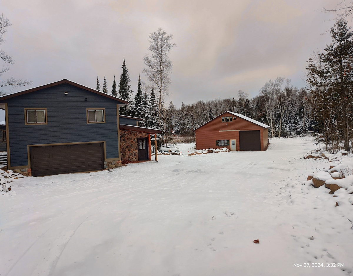 A spacious exterior view showcases a two-story house alongside a separate building, both surrounded by snow-covered ground. Evergreen trees border the scene, while a cloudy sky adds a soft light to the tranquil winter landscape.