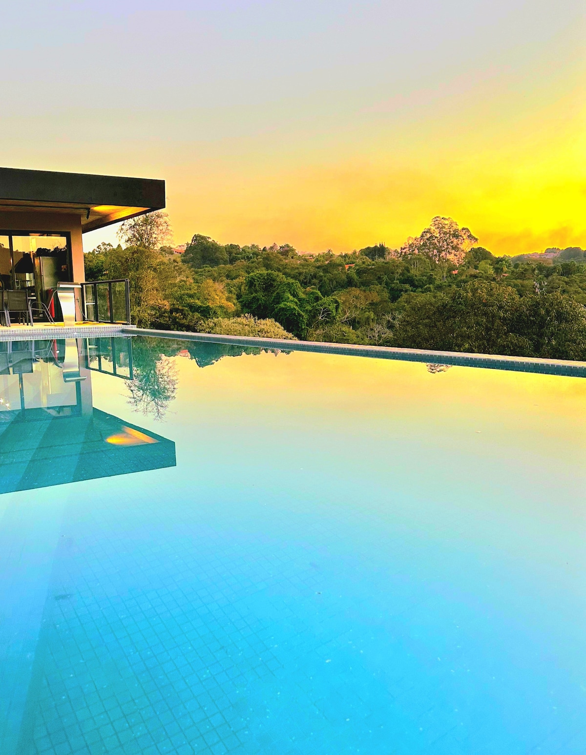 An infinity pool reflects the serene colors of the sunset, with lush green landscapes visible in the background. The pool's smooth surface creates a seamless connection with nature, while nearby, glass doors lead into the interior of the house.